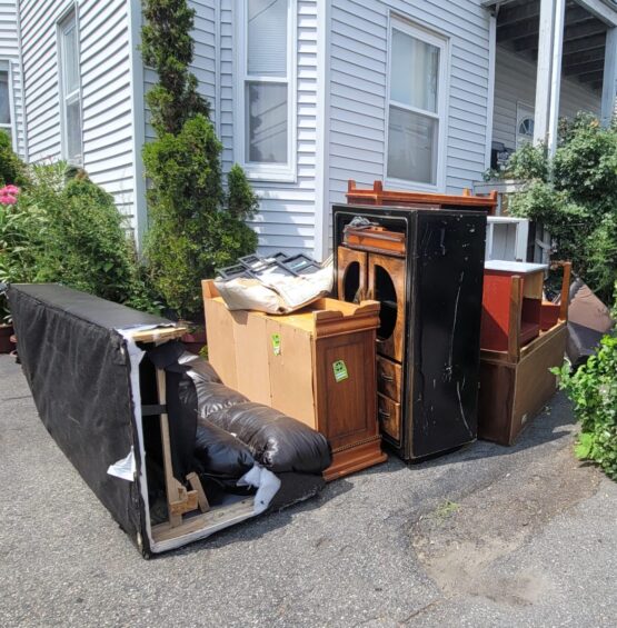 A pile of discarded furniture and household items, including a couch, a broken dresser, and a disassembled shelving unit, left on a sidewalk next to a residential street on a sunny day.
