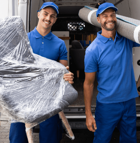 Two smiling movers in blue uniforms carrying a wrapped armchair and a rolled-up carpet, ready to load into their junk removal truck.